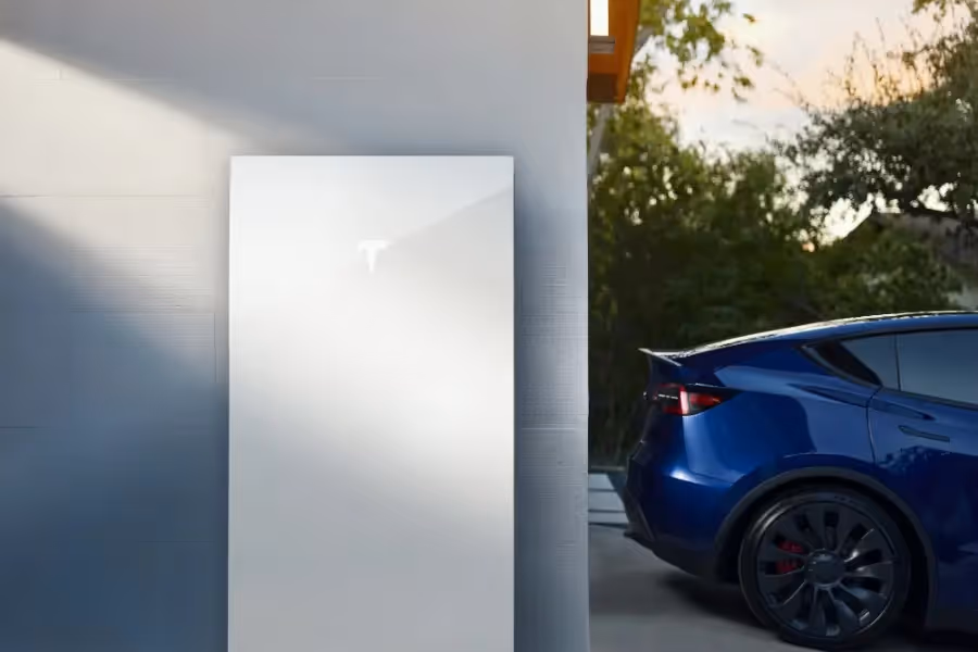 A Tesla Powerwall 3 on a wall with a Tesla car on the driveway behind