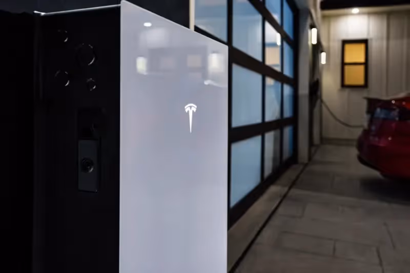 A Tesla Powerwall 3 on the wall outside a house, with a Tesla car in the background