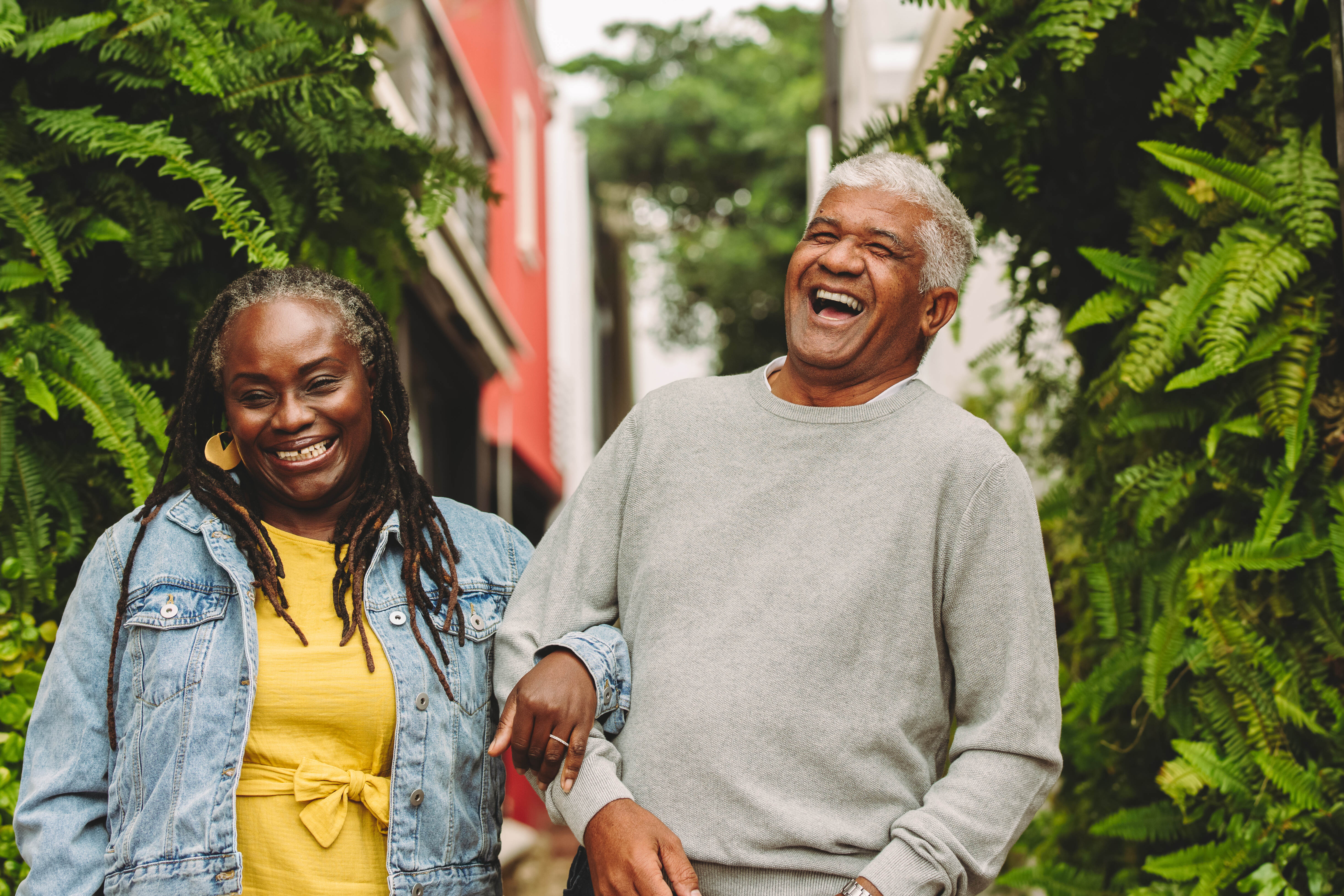 Smiling elderly couple walking arm in arm outdoors with greenery and buildings in the background.