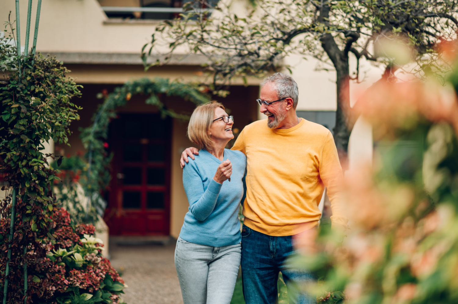 Smiling older couple, woman in blue sweater and man in yellow sweater, walking arm in arm outdoors near a garden.
