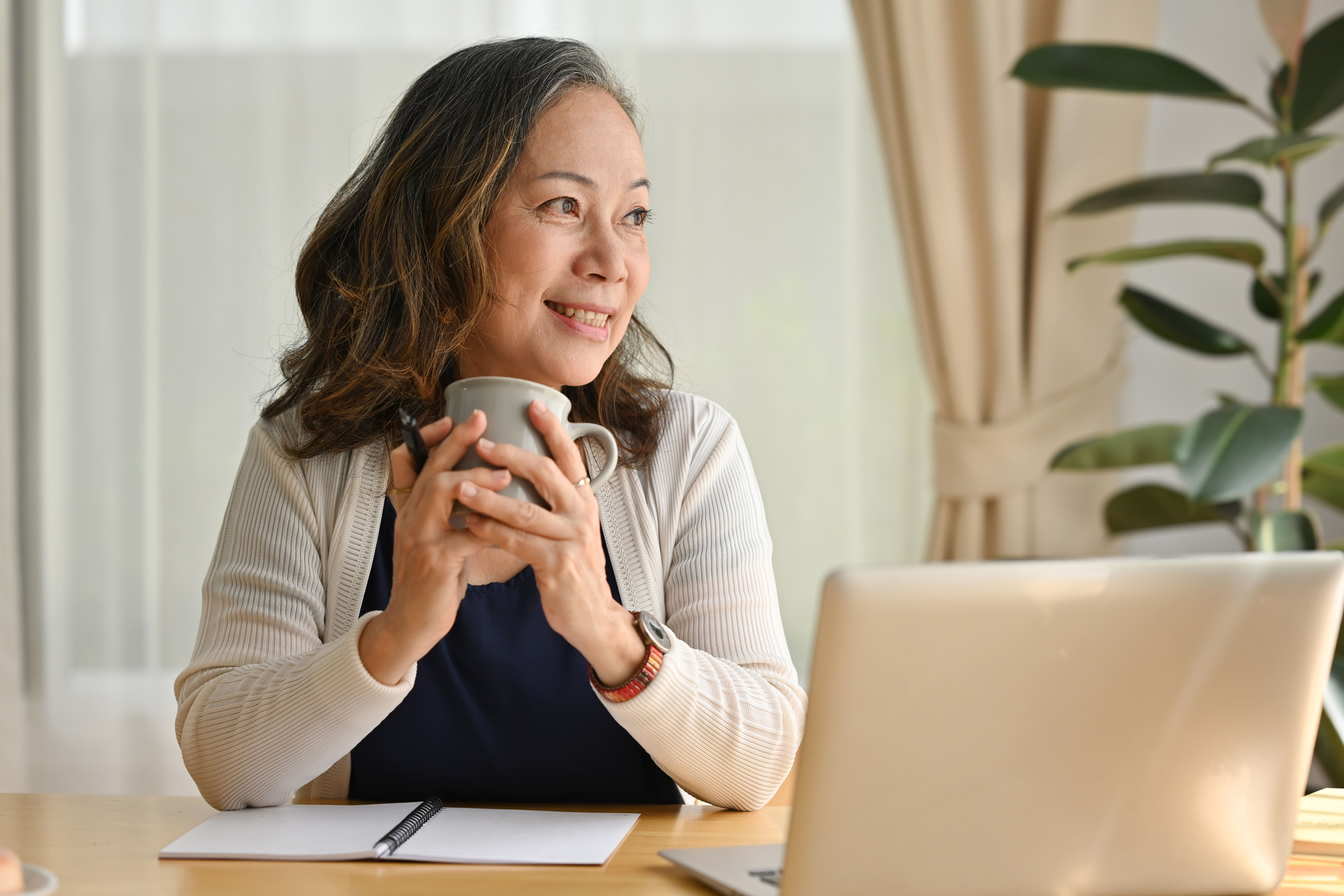 Smiling woman holding a mug and looking thoughtfully to the side at a desk with a laptop and notebook.