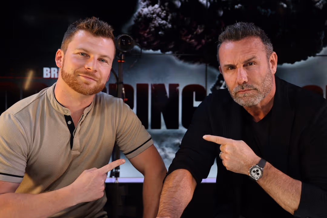 Two men sitting side by side, each pointing at the other with a serious expression, against a dark background with partially visible text.