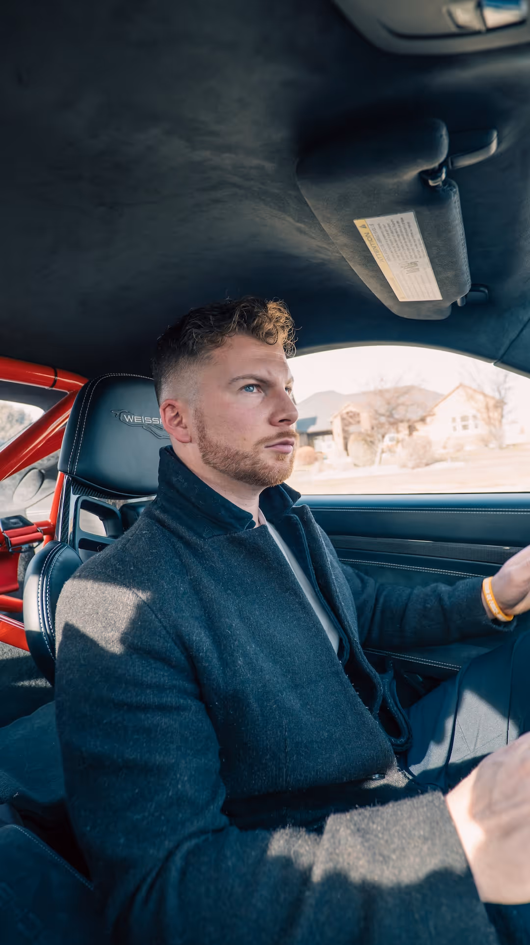 Man with short curly hair and beard driving a car with a Weissach seat and visible red roll cage.