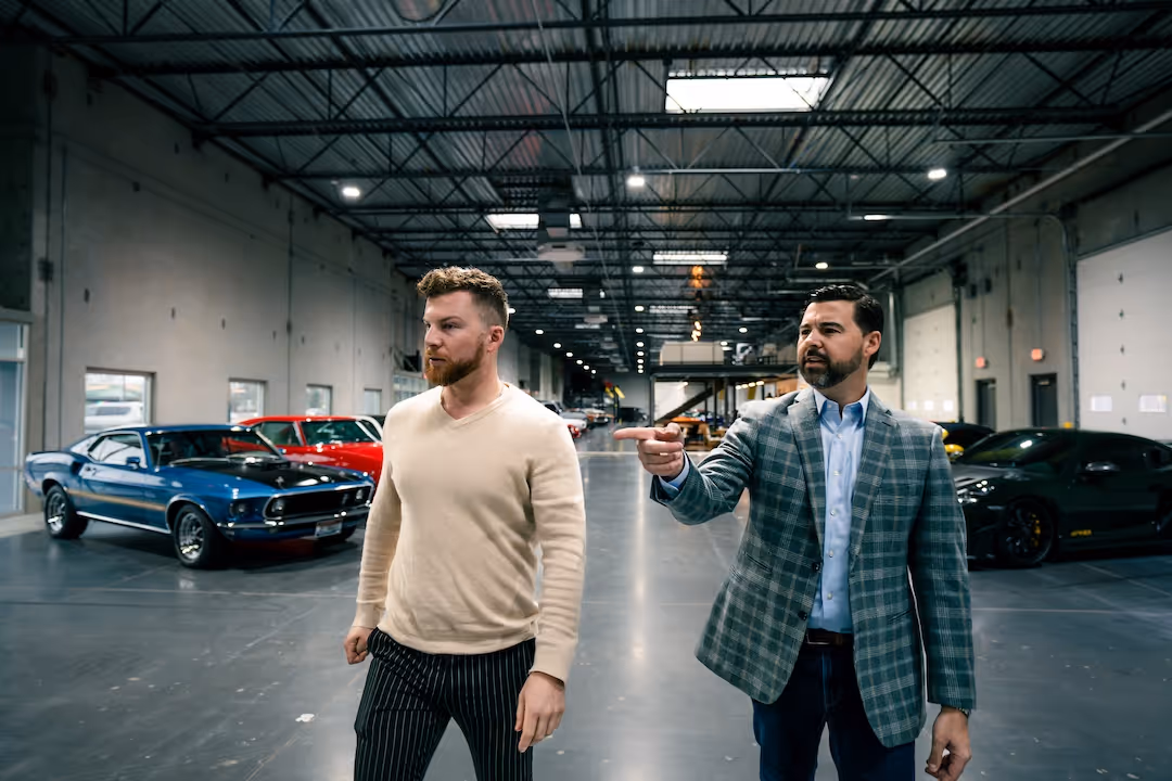 Two men standing in a large garage with classic cars in the background, one man pointing to the side.