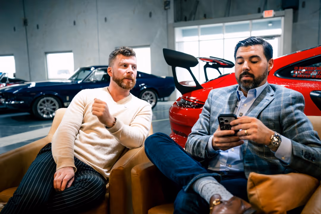Two men sitting in armchairs in a garage with luxury cars in the background; one man is looking at the other while the other checks his smartphone.