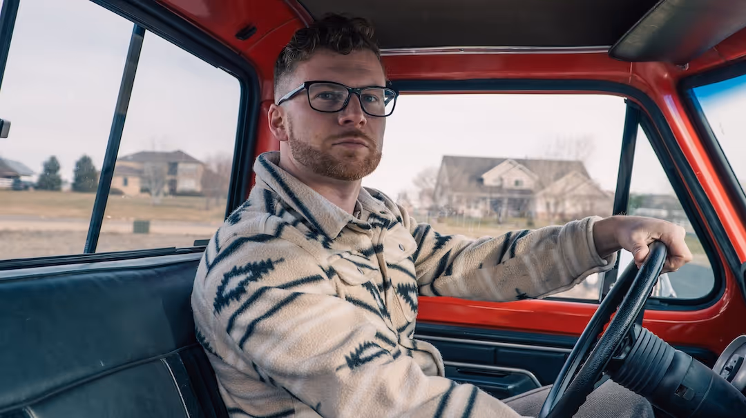 Man wearing glasses and a patterned jacket driving a red vintage truck near suburban houses.