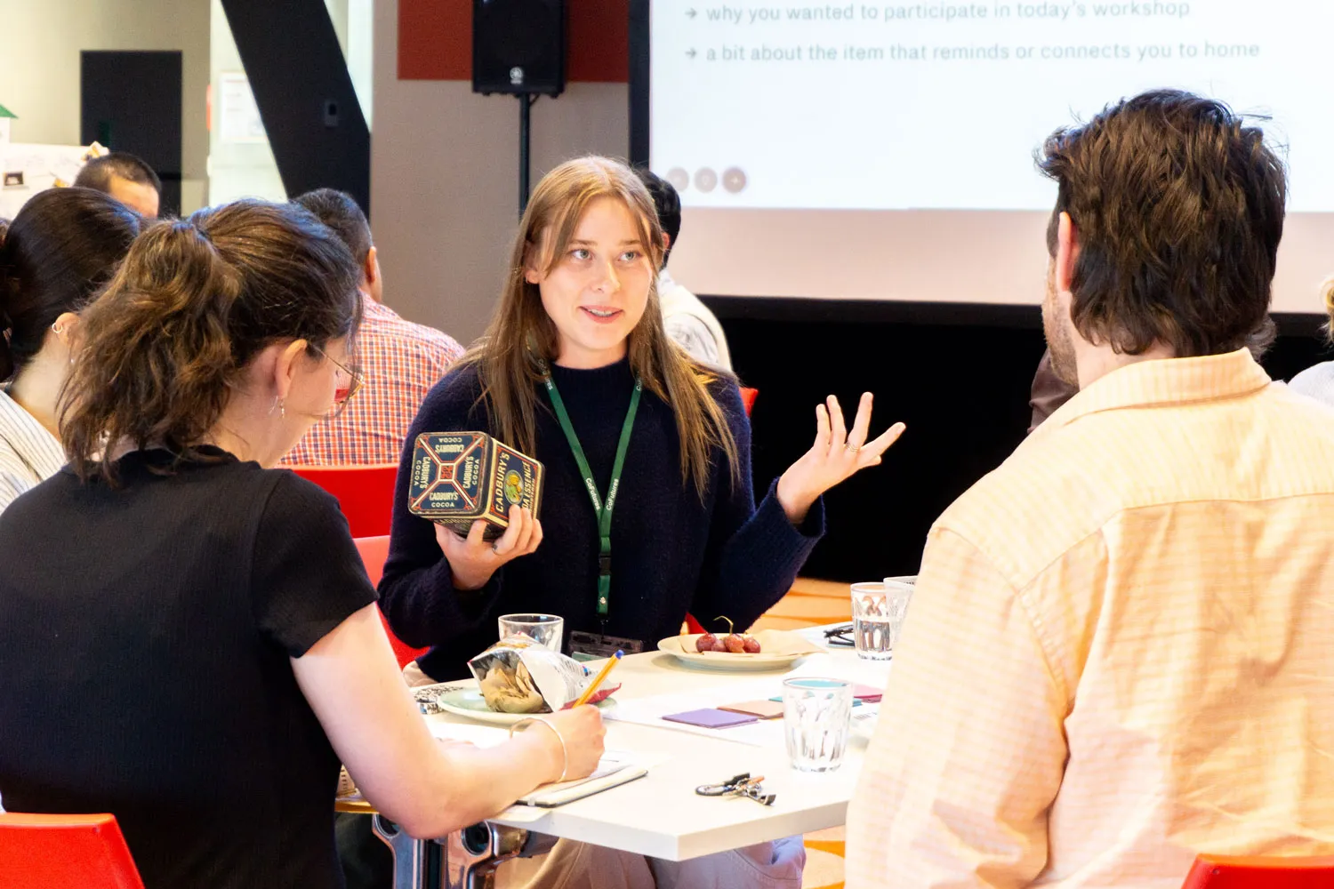 Woman discussing and holding a vintage Cadbury cocoa tin while seated at a table with others in a workshop setting.