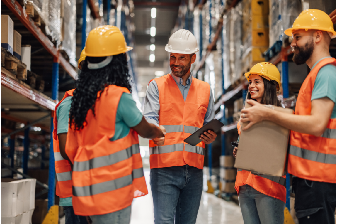 Warehouse workers wearing orange safety vests and helmets shaking hands and holding packages in an aisle with stacked shelves.