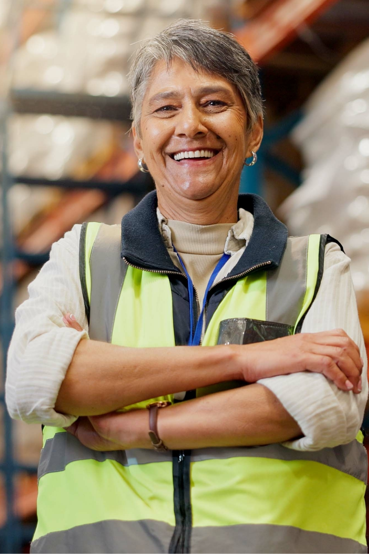 Smiling middle-aged woman wearing a high-visibility safety vest with arms crossed in a warehouse.