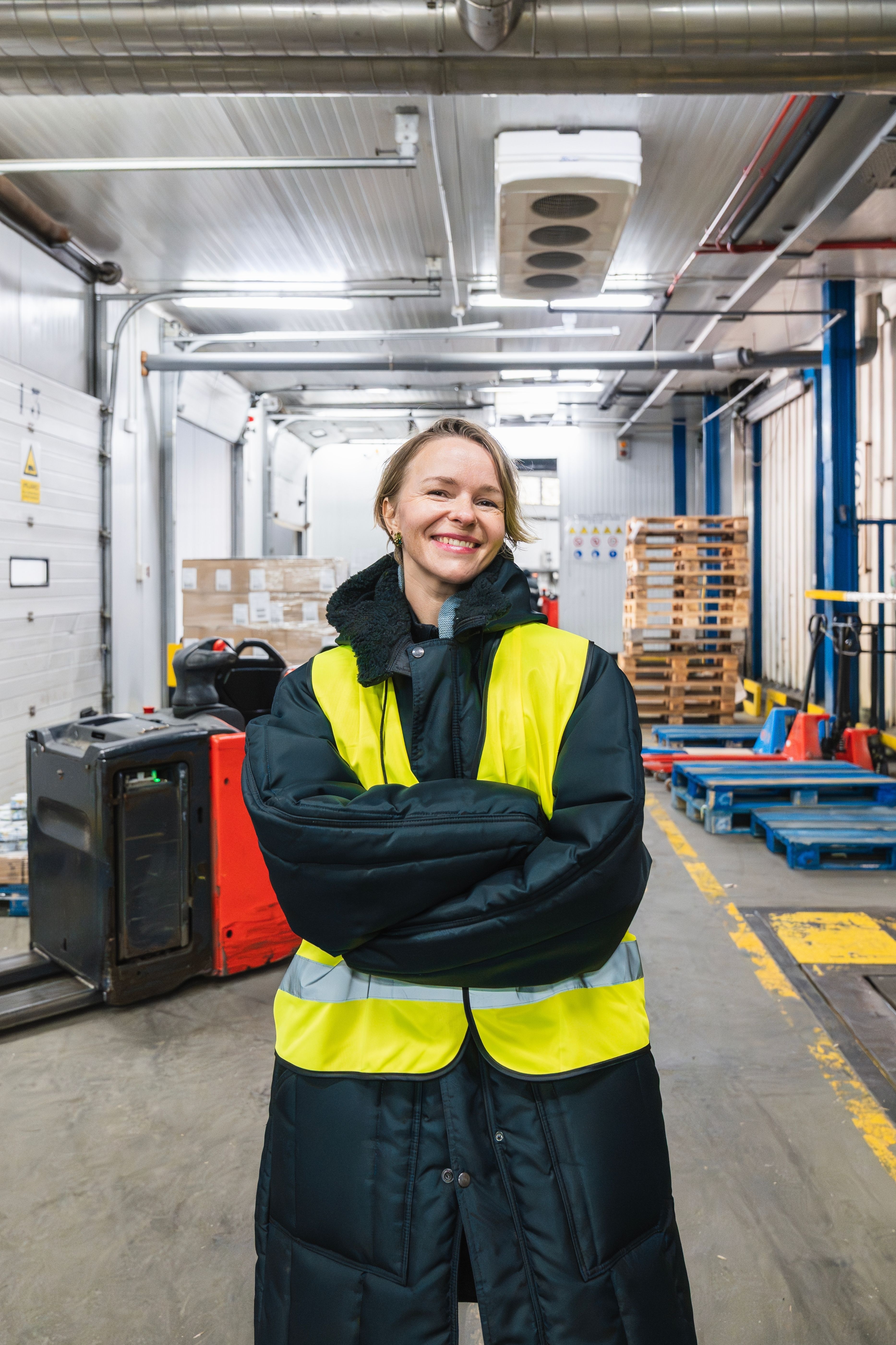 Smiling woman wearing a yellow safety vest and black winter coat standing with arms crossed in a warehouse.