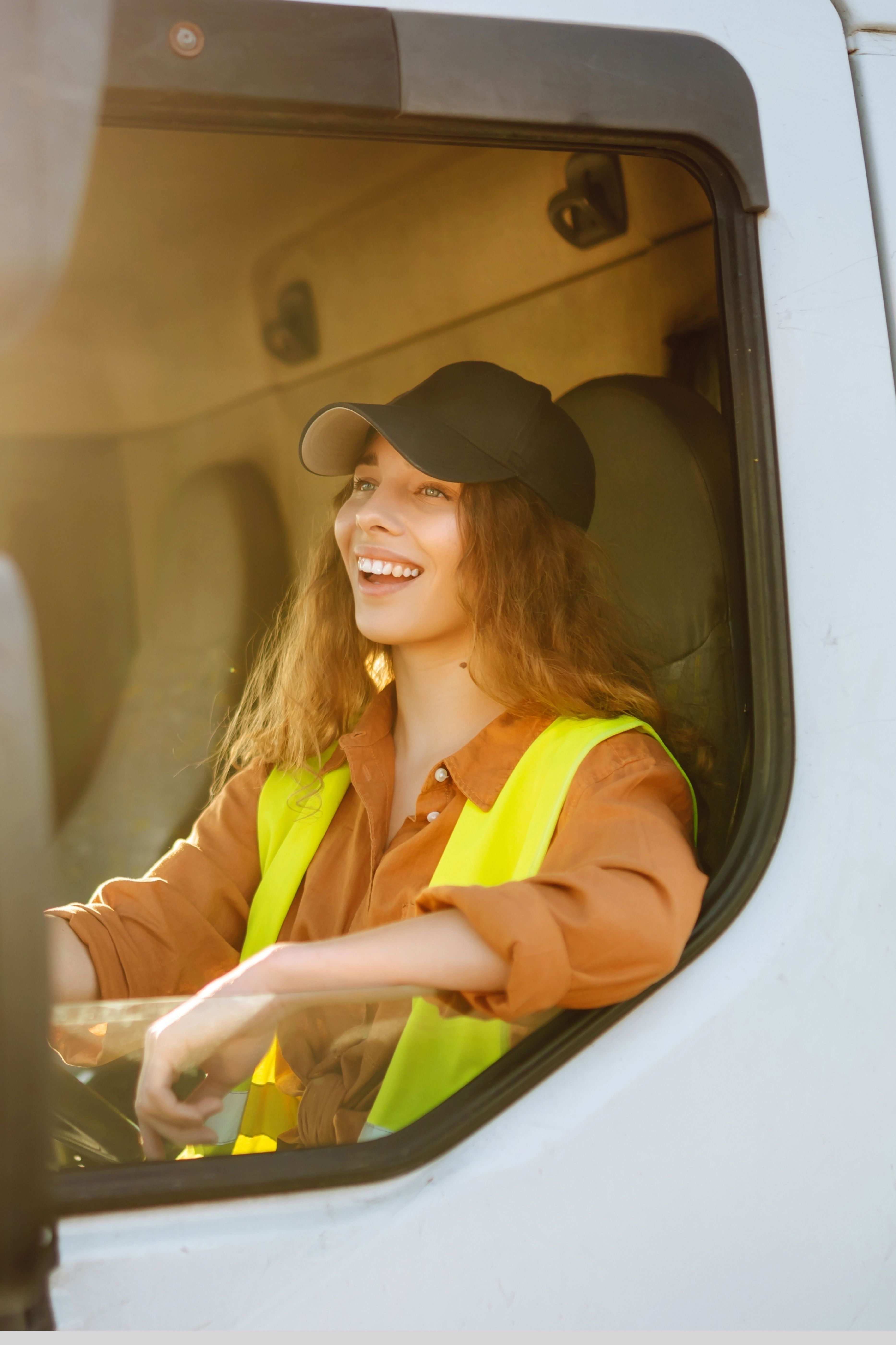 Smiling woman wearing a black cap and yellow safety vest sitting in the driver's seat of a white vehicle.
