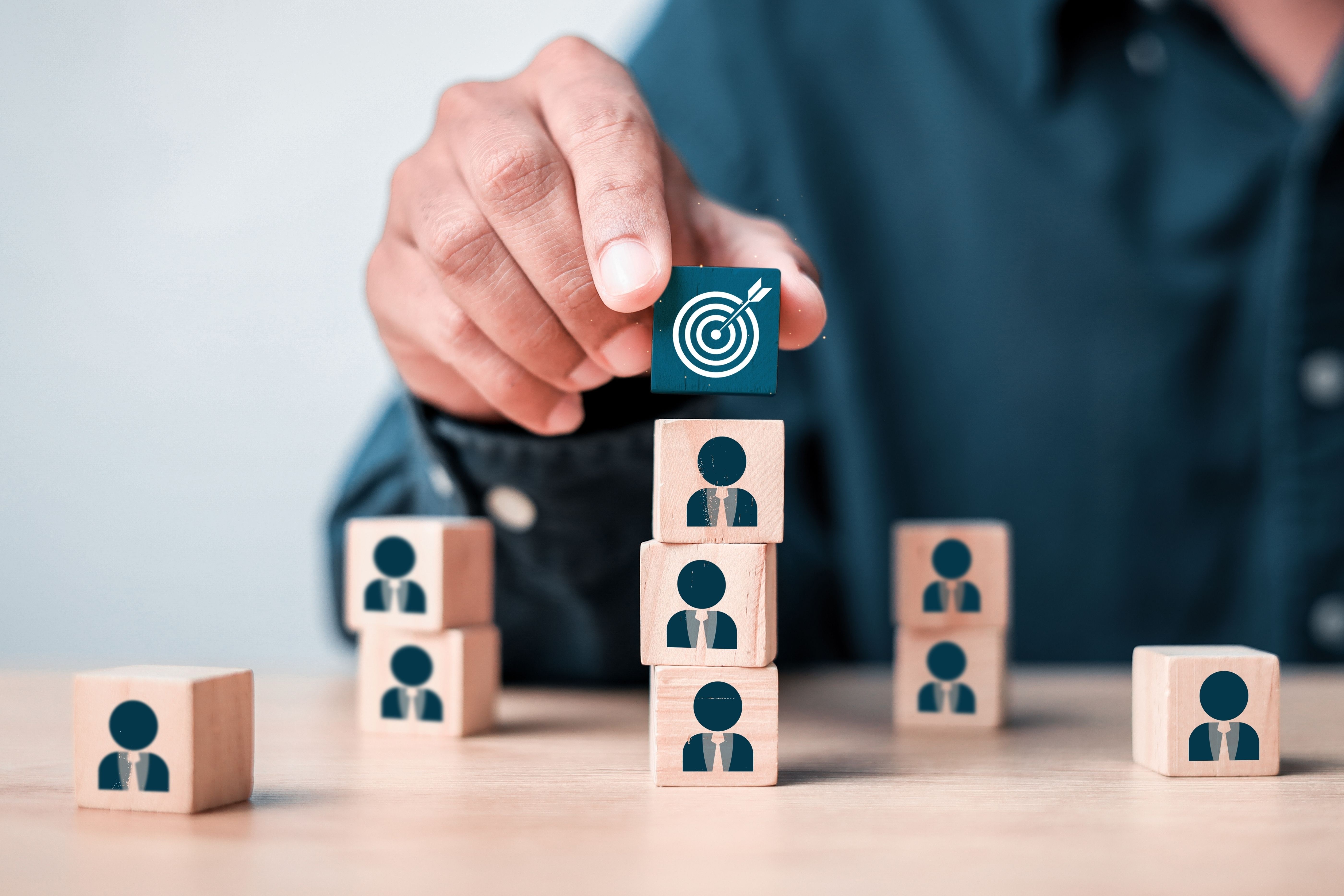 Hand placing a block with a target icon on top of stacked wooden blocks with person icons.