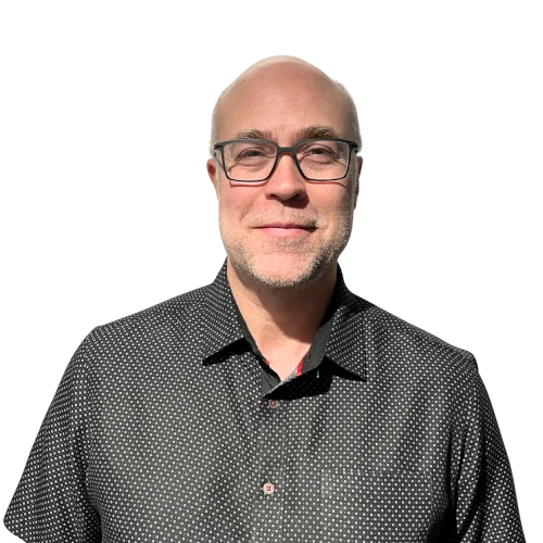 Homme chauve portant des lunettes et une chemise noire à petits motifs blancs, souriant légèrement devant un fond transparent.