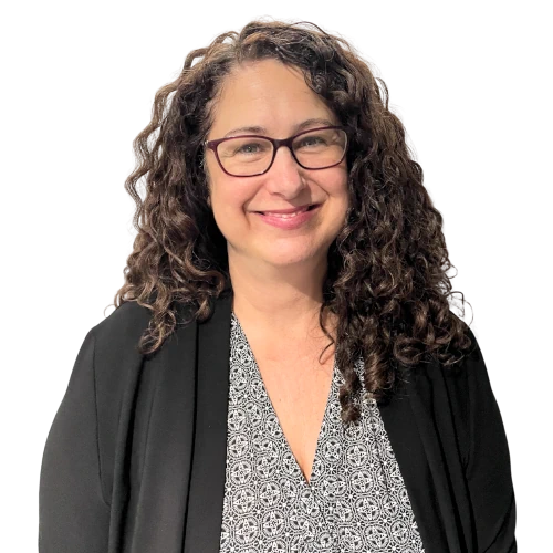 Femme souriante avec des cheveux bouclés bruns, portant des lunettes et un blazer noir sur un haut à motif blanc et noir.