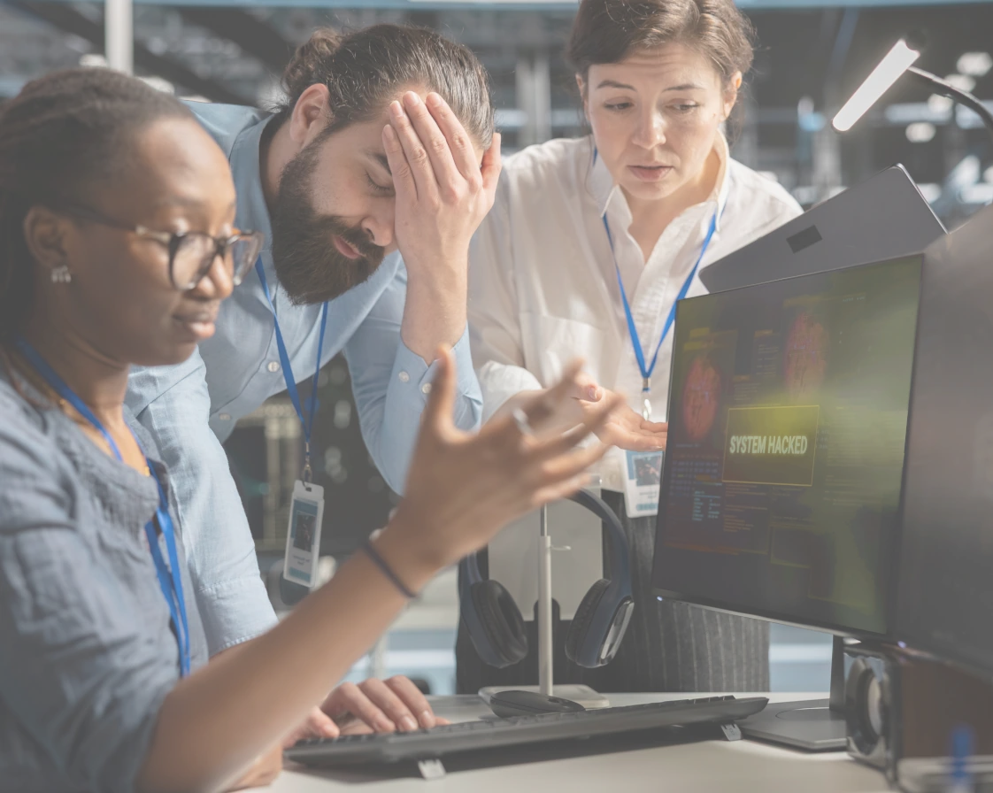 Trois professionnels informatiques préoccupés devant un écran d'ordinateur affichant le message 'SYSTEM HACKED'.