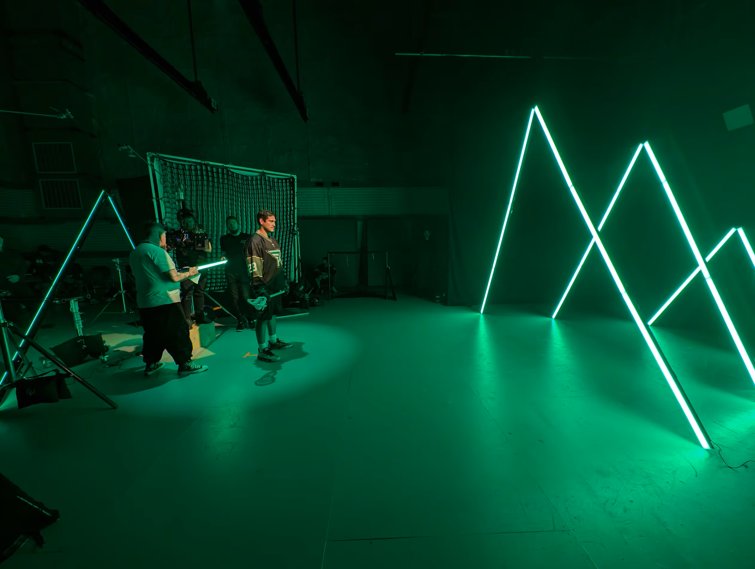 Man in hockey gear stands near net and crew holding green light tubes in dark room with green neon triangles.