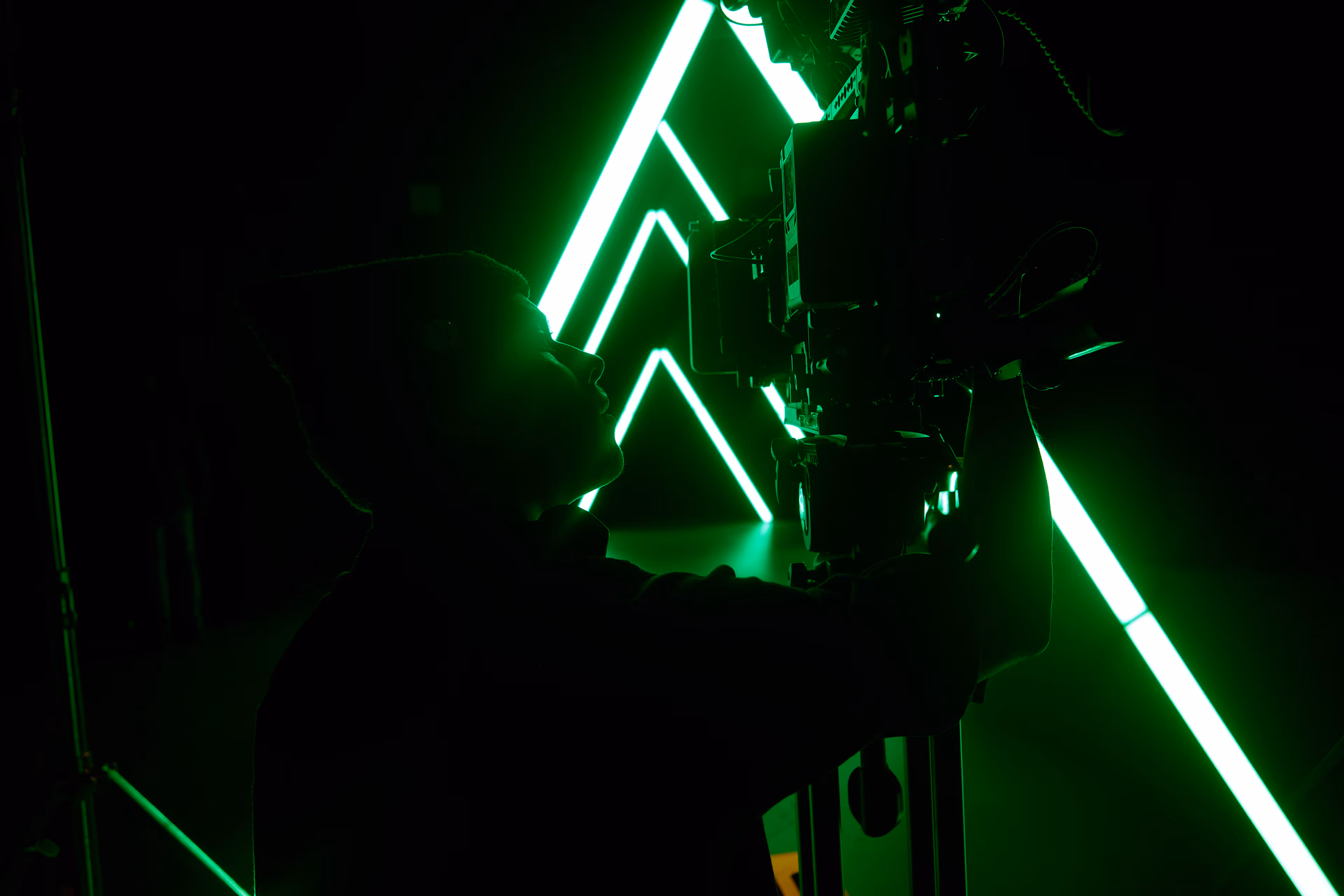 Silhouette of a person operating a camera with green neon triangular lights in the background.