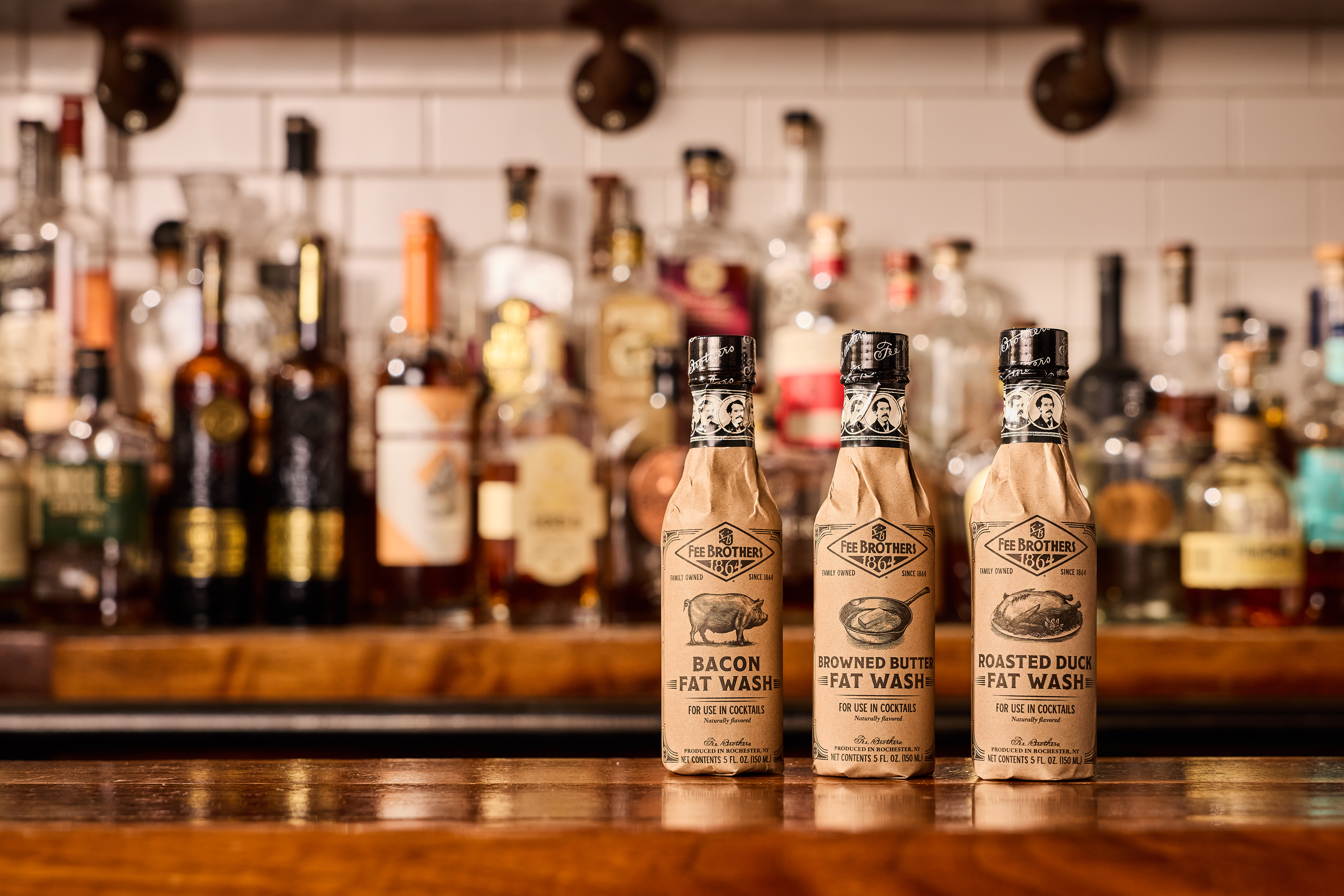 Three Fee Brothers Fat Wash bottles wrapped in brown paper on a bar with liquor bottles blurred behind.