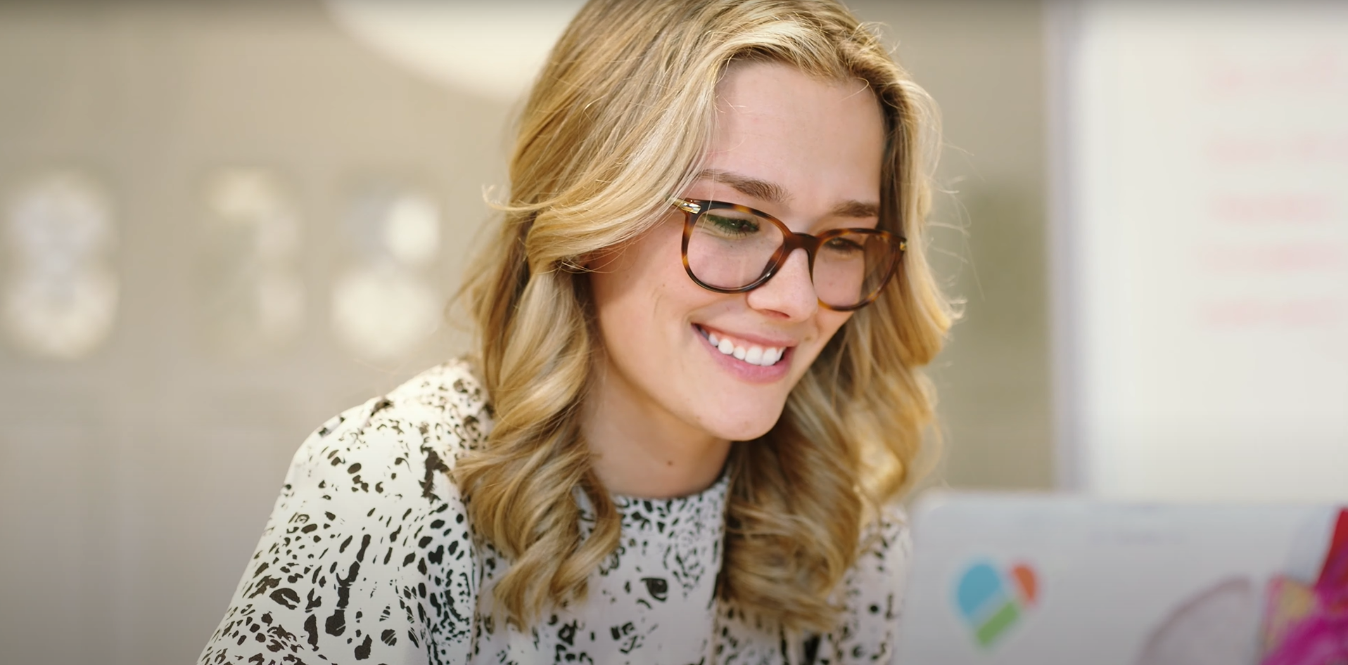 Smiling woman with glasses and blonde curly hair looking at a laptop with a colorful sticker.