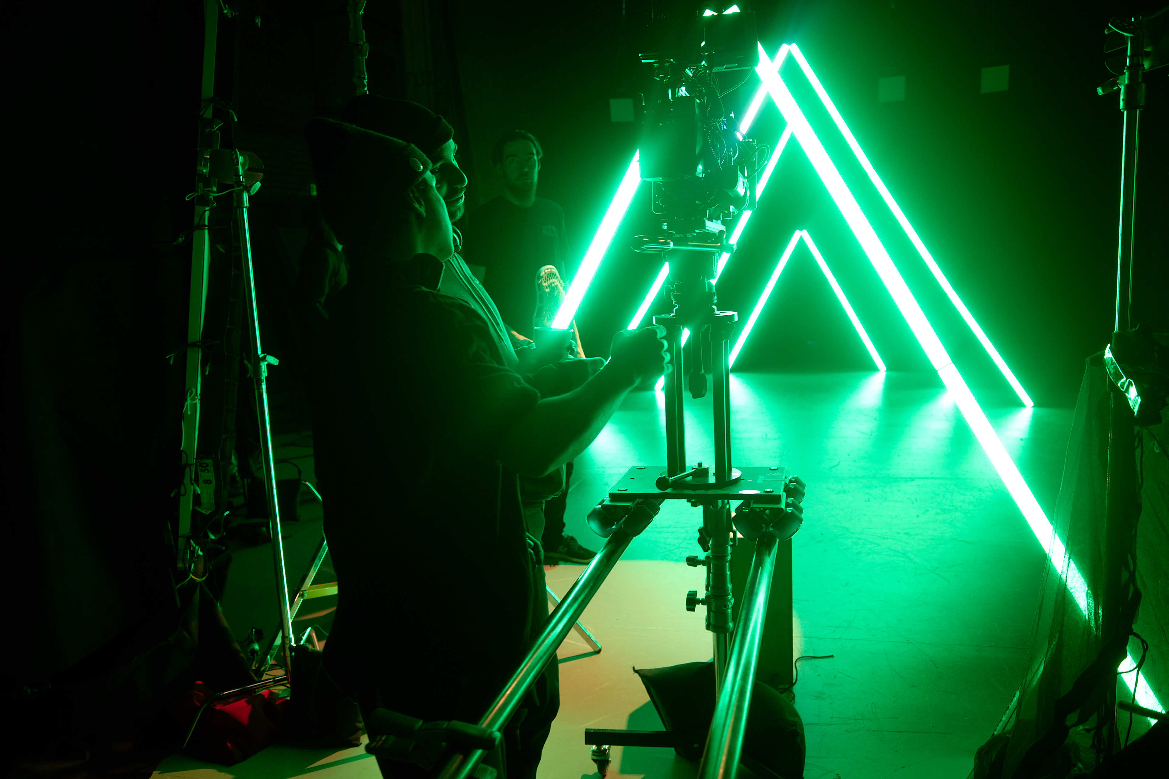 Three men operating a camera on a dolly rig with bright green neon triangular lights in the background.