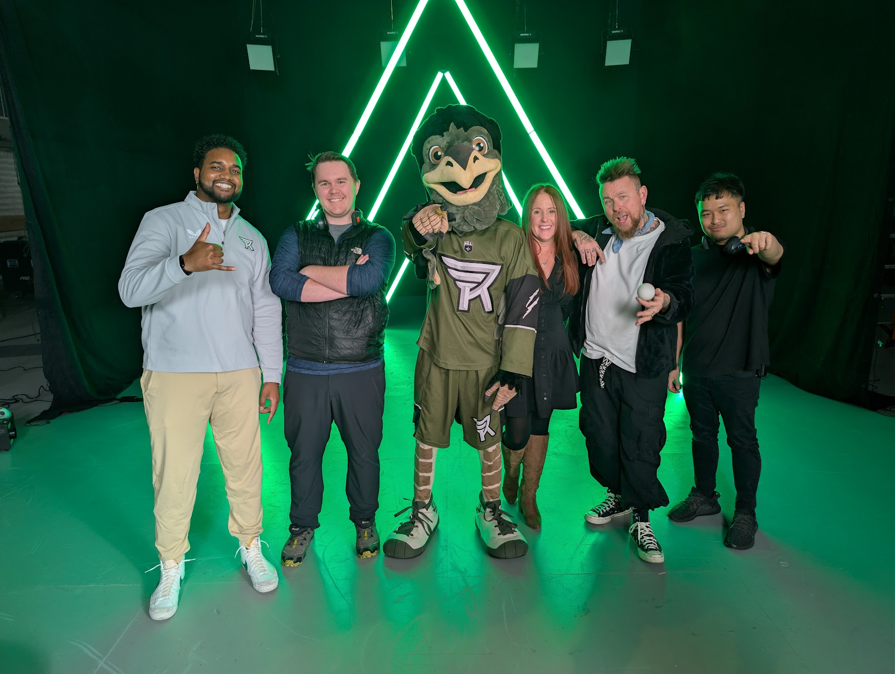 Five people and a bird mascot stand smiling in front of green neon lights shaped like triangles.