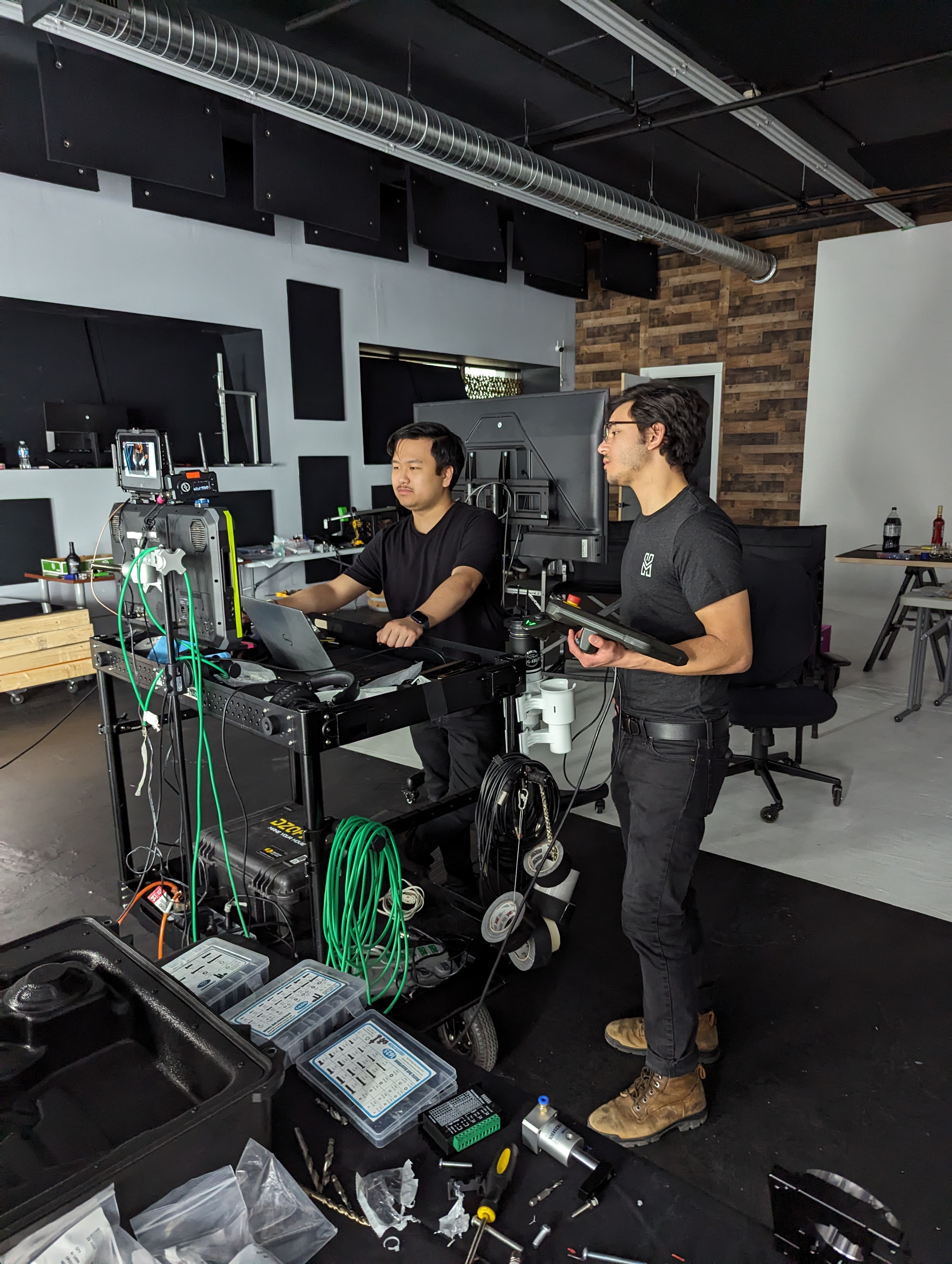 Two men working with video and computer equipment on a cart in a studio with tools and cables nearby.