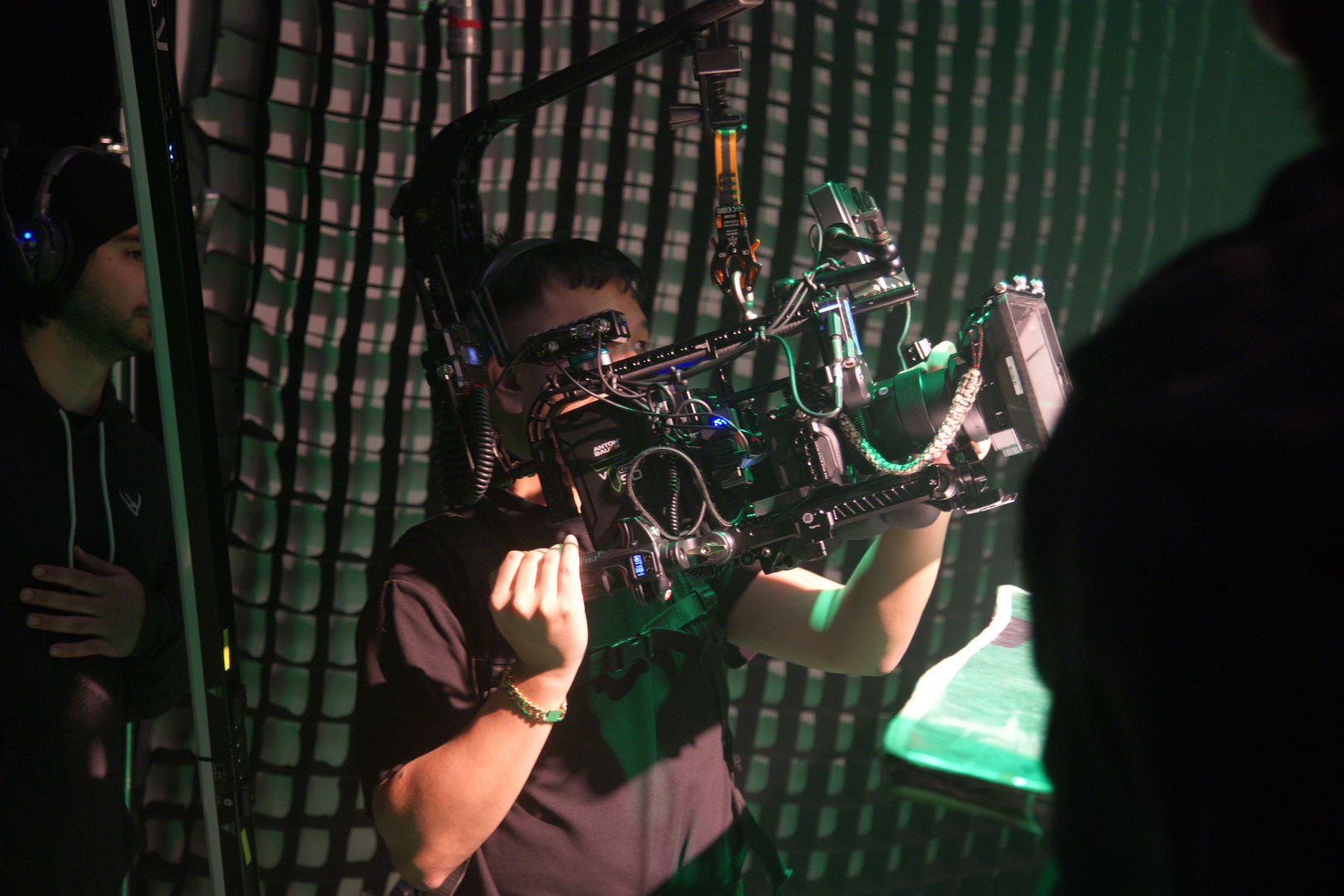 Person operating a complex camera rig with mounted monitors and cables indoors, another person nearby.