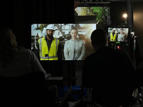 Two people watching a video screen showing a man in a safety vest and a woman in a gray jacket.