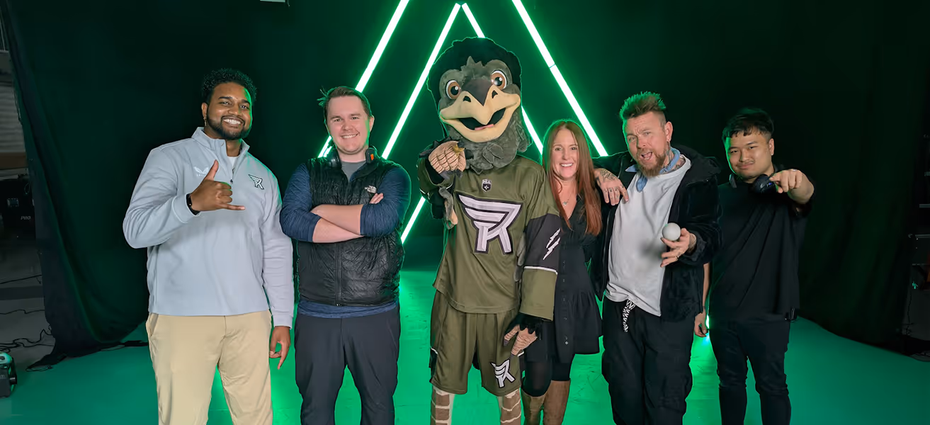 Six people, including a person in a bird mascot costume, posing with a neon green triangular light background.