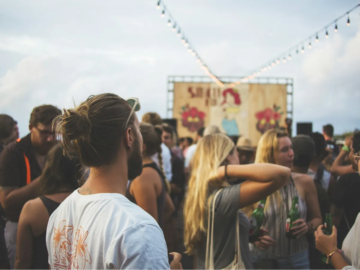 Crowd of people socializing at an outdoor event with string lights overhead and a colorful mural in the background.
