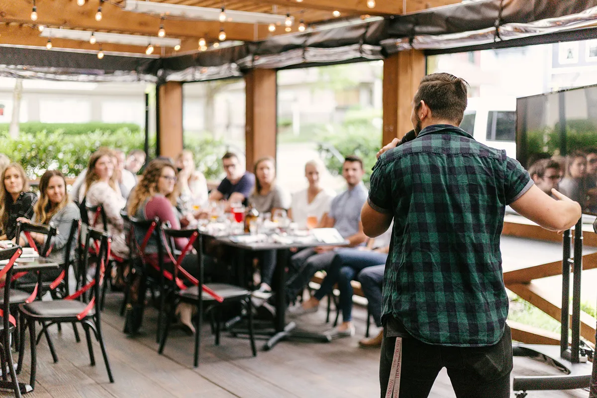 Man speaking with a microphone to an audience seated around tables in an outdoor covered patio.