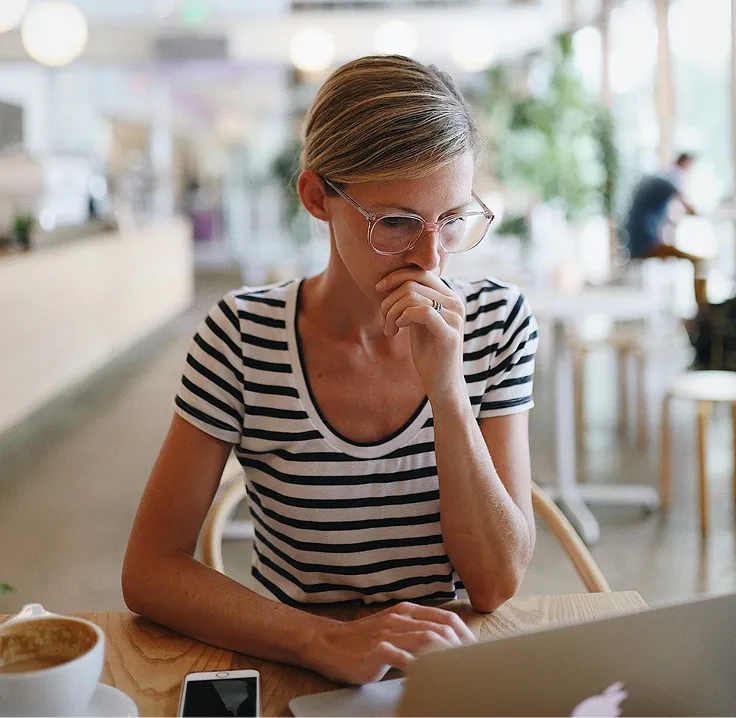 Woman with glasses wearing a striped shirt working on a laptop in a bright café.