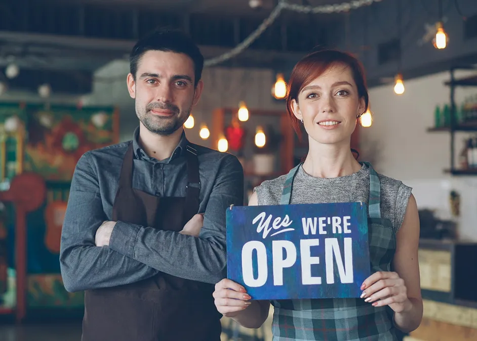 Smiling man and woman wearing aprons, with the woman holding a sign that says 'Yes we're open' in a warmly lit shop.