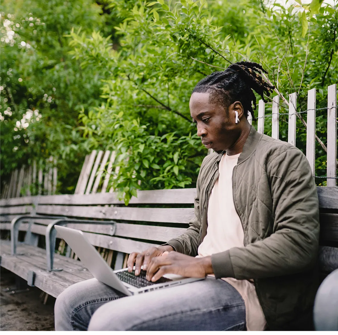 Young man with dreadlocks sitting on a wooden bench outdoors typing on a laptop, with green foliage in the background.