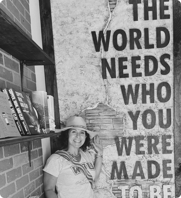 Smiling woman wearing a hat and t-shirt standing next to a wall with large text reading 'The world needs who you were made to be'.