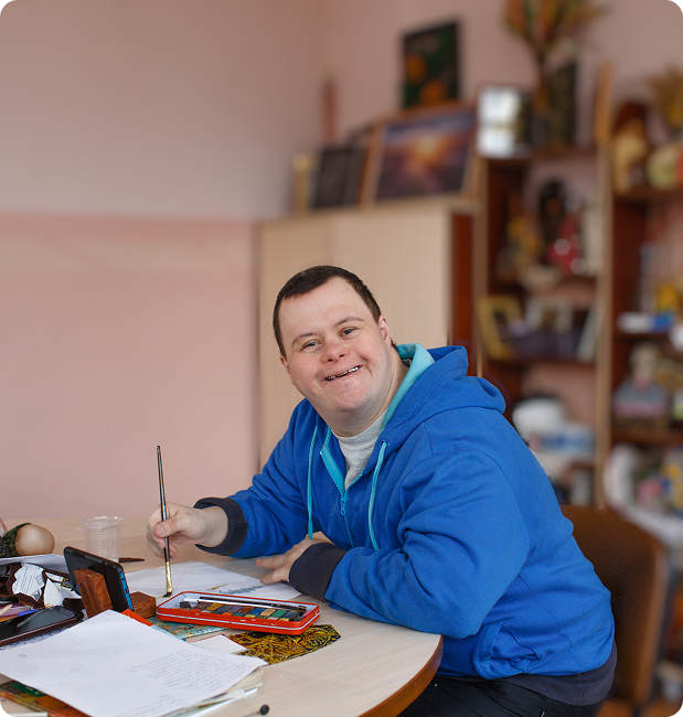 Smiling man in a blue hoodie sitting at a table with watercolor paints and a brush, preparing to paint.