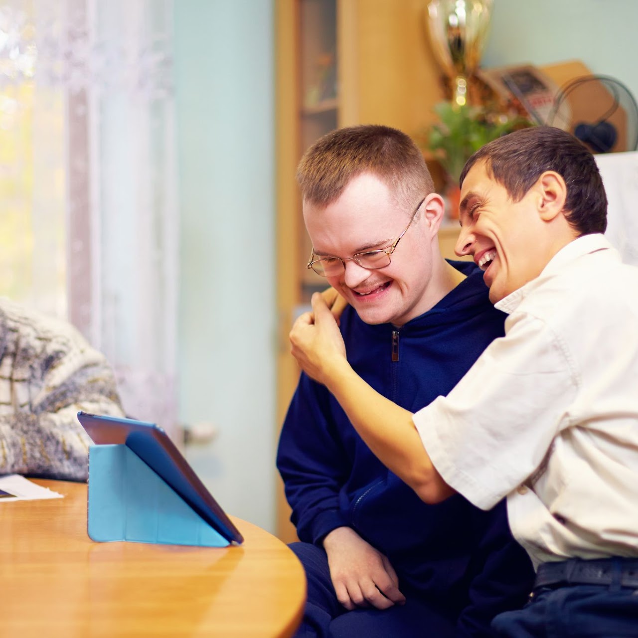Two men smiling and laughing together while looking at a tablet on a wooden table indoors.