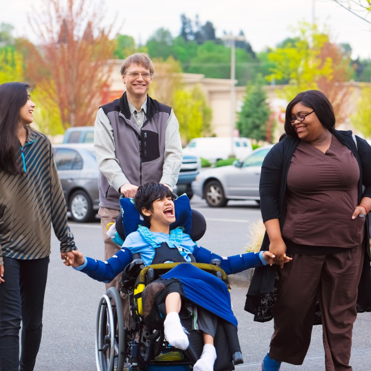 Smiling young person in a wheelchair holding hands with two women while a man pushes the wheelchair outside near parked cars.