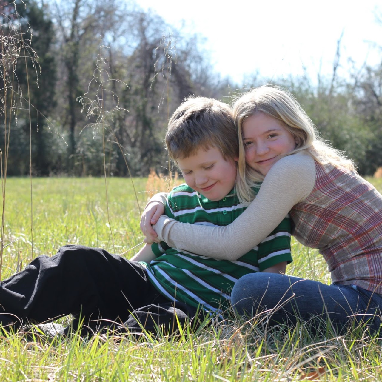 Young girl hugging a smiling boy while sitting together on grass in a sunny outdoor setting.