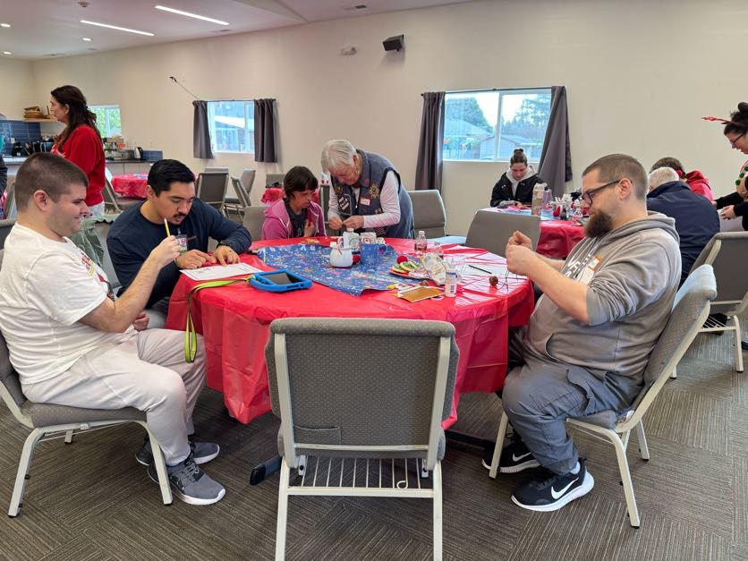 Group of people engaged in crafting activities while sitting around a table covered with a red tablecloth and crafting materials in a bright room.