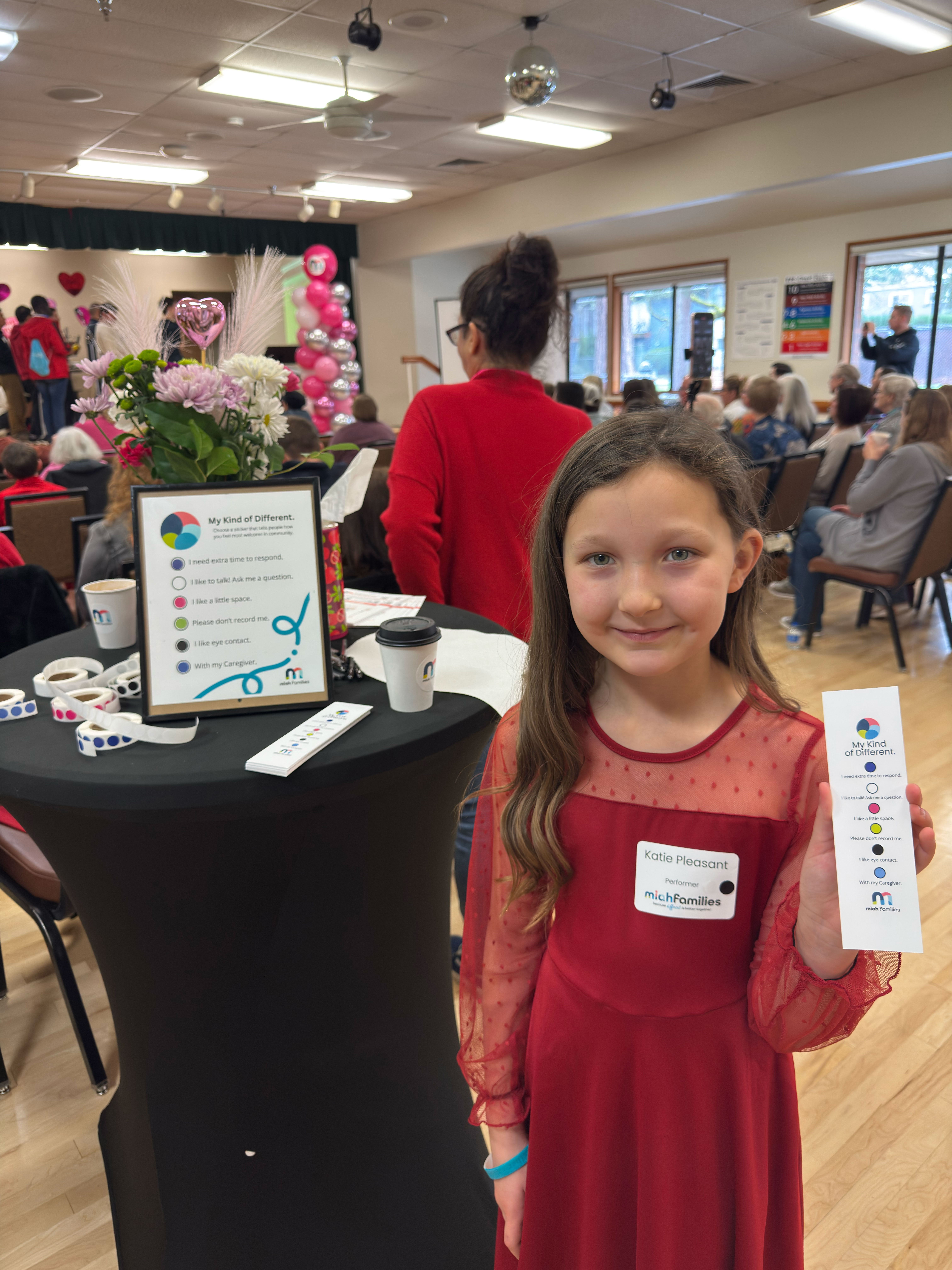 Young girl in a red dress with a name tag reading 'Katie Pleasant Performer' holding a 'My Kind of Different' card with communication preferences in a community event room.
