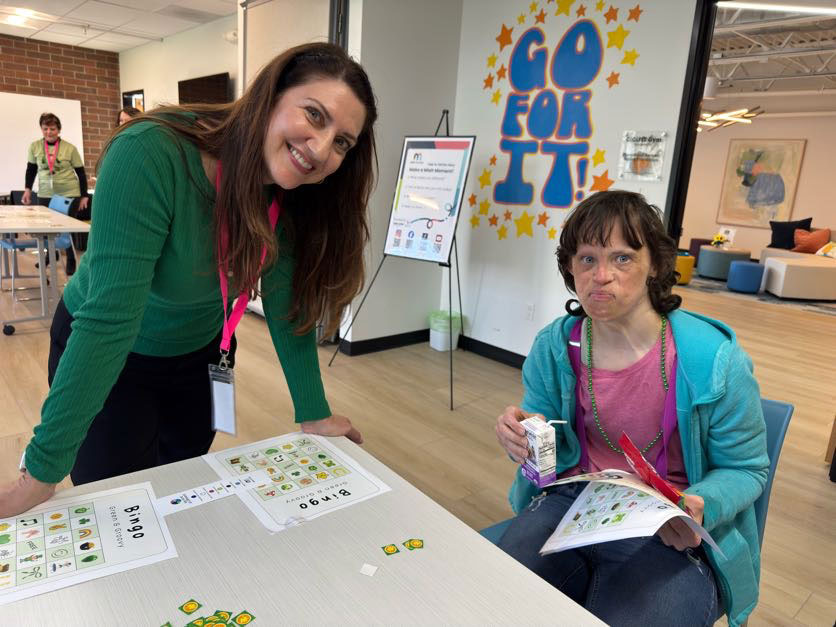 Two women playing bingo at a table in a community center with a 'GO FOR IT!' sign on the wall behind them.