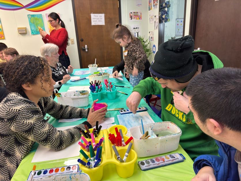 A group of people of various ages painting and drawing together at a table with art supplies and watercolors.