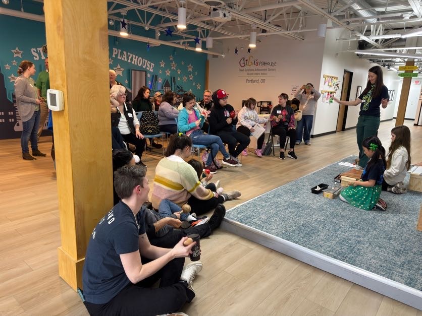 Group of adults and children seated and listening to a woman leading an activity on a raised carpeted platform in a playhouse room.