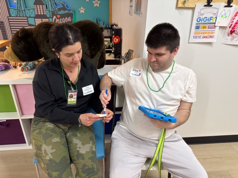 A woman assisting a man with a blue marker, both wearing name tags and green bead necklaces, sitting in a colorful room with a large stuffed bear in the background.