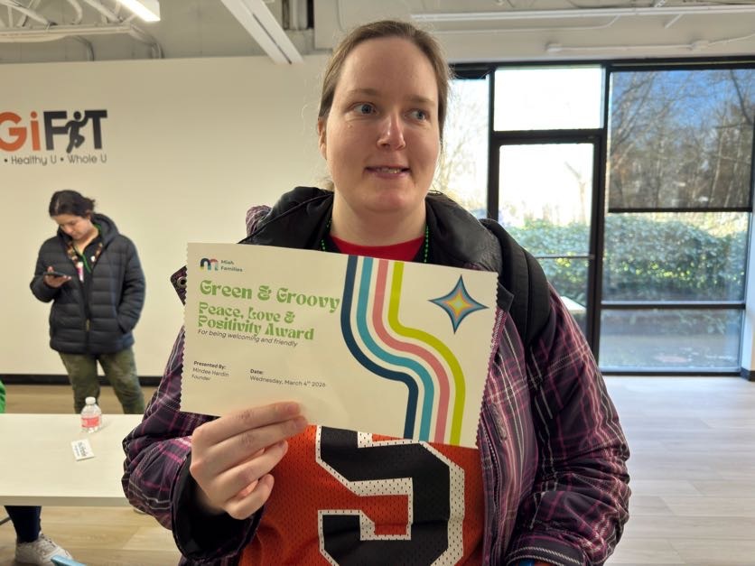 Woman holding a Green & Groovy Peace, Love & Positivity Award certificate inside a room with large windows.