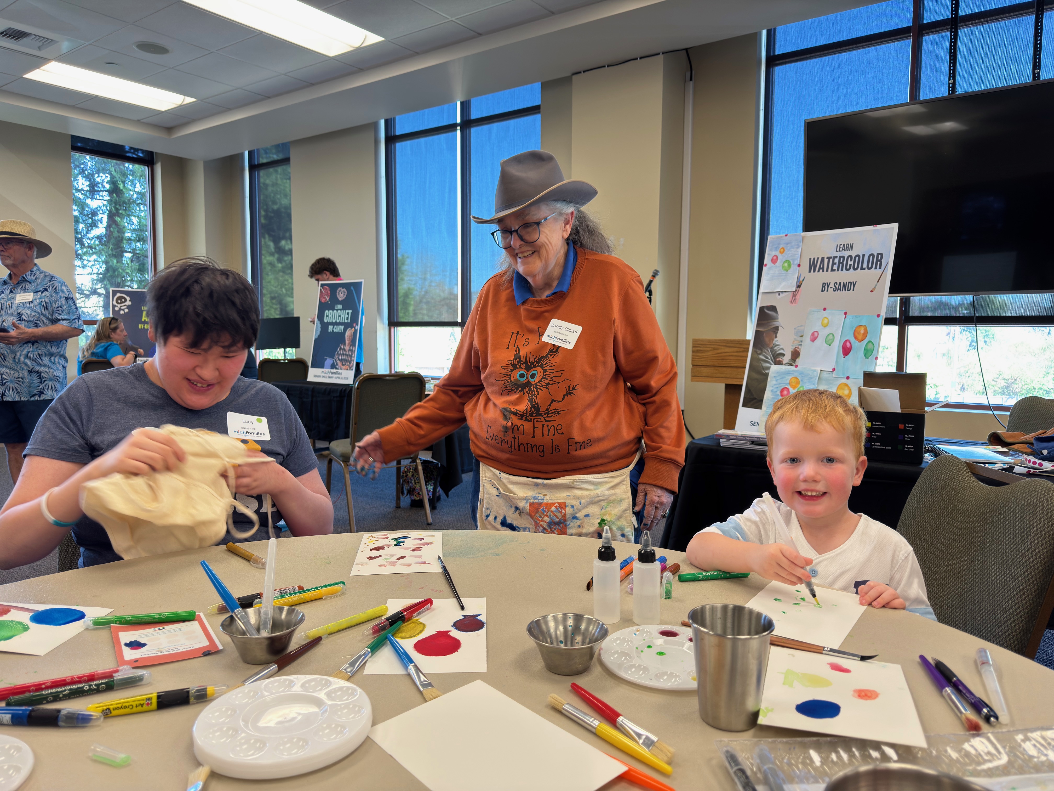 A person in a yellow Minion hat and shirt helps a boy in an orange jacket with a craft involving black paint and a sponge on an orange tablecloth.