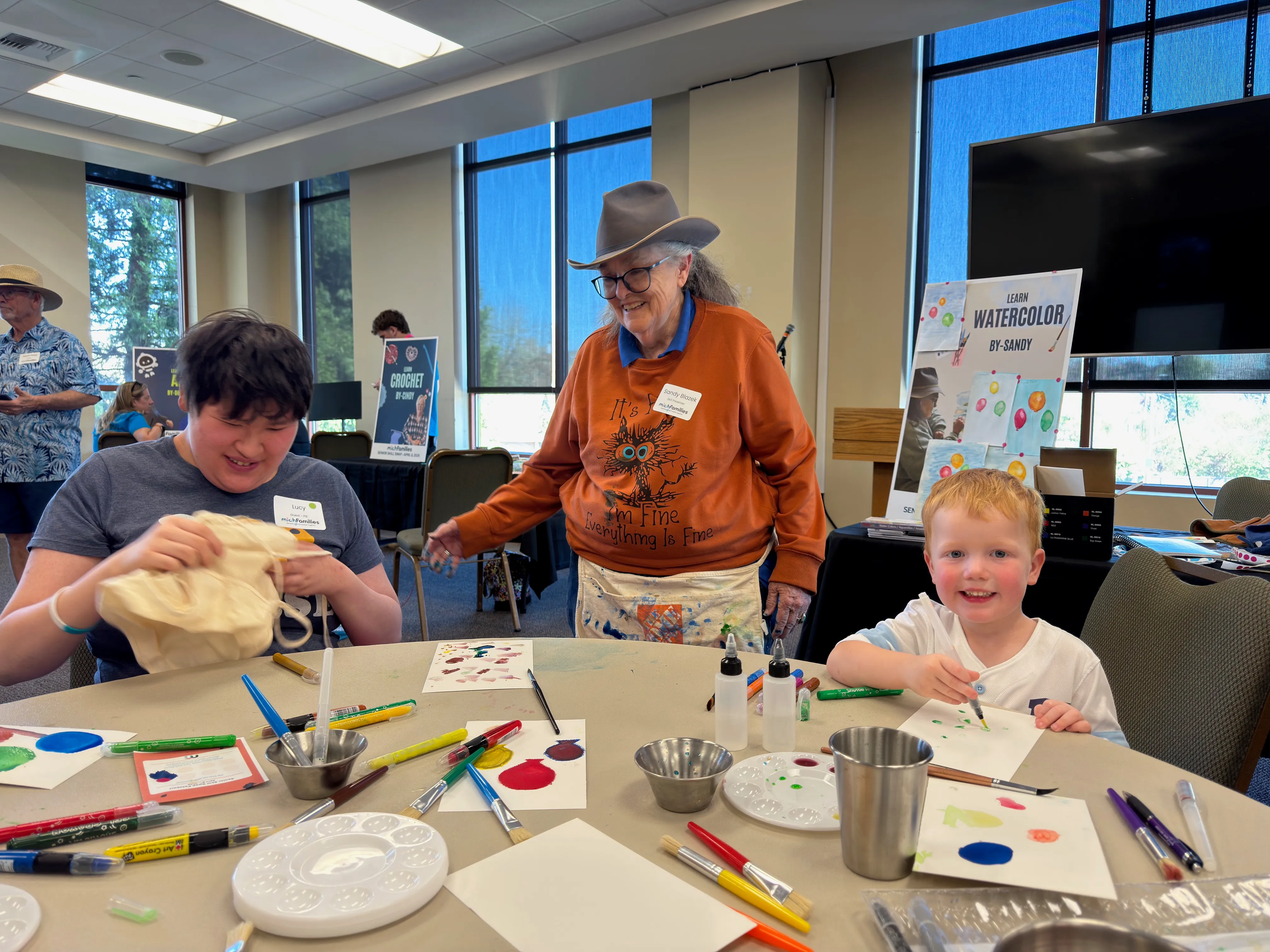 A person in a yellow Minion hat and shirt helps a boy in an orange jacket with a craft involving black paint and a sponge on an orange tablecloth.