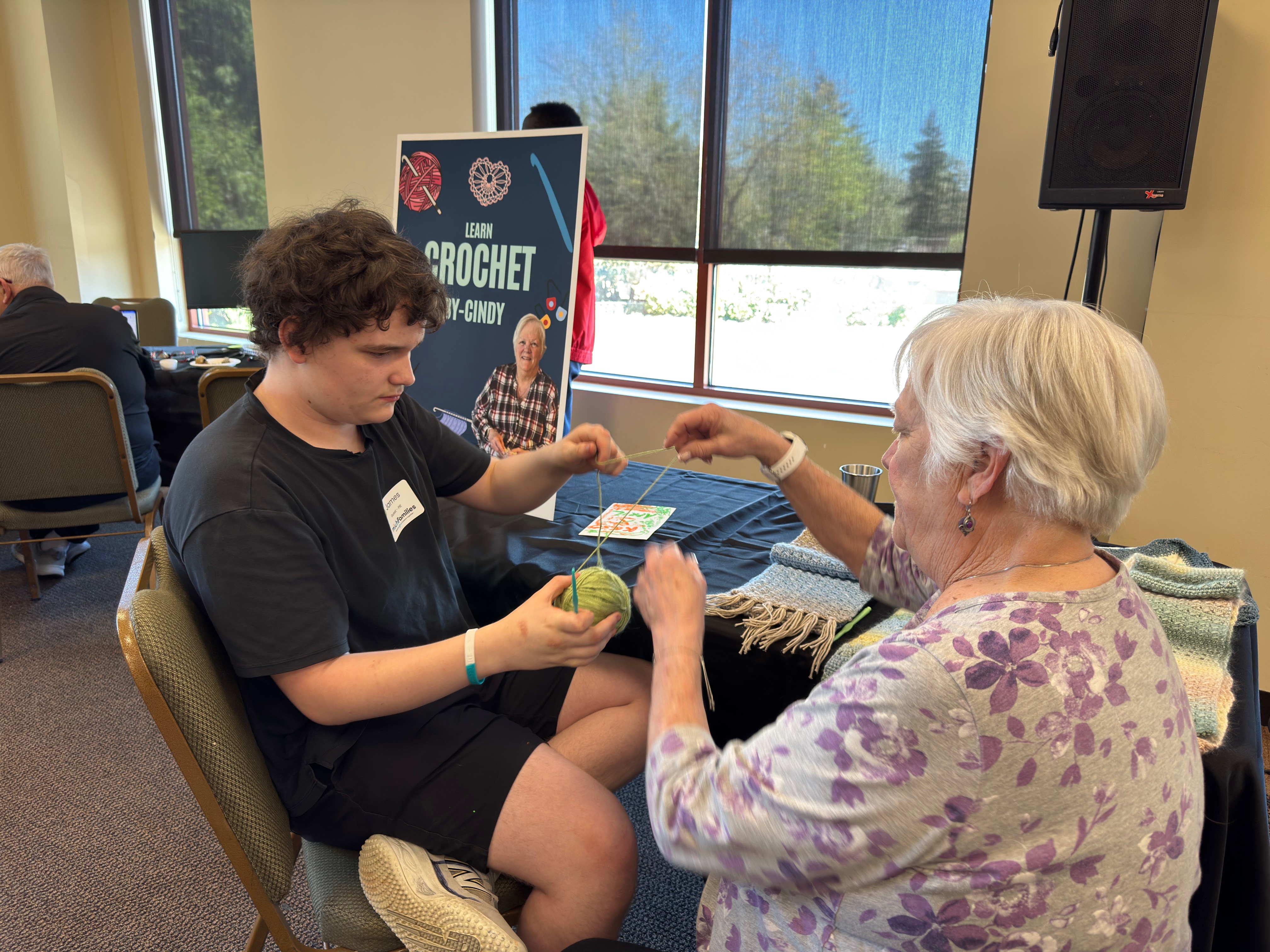 Woman in a yellow polka dot jacket and pink checkered hat feeding a whipped cream-covered treat to a young girl in a pink and white outfit at a table with autumn decorations indoors.
