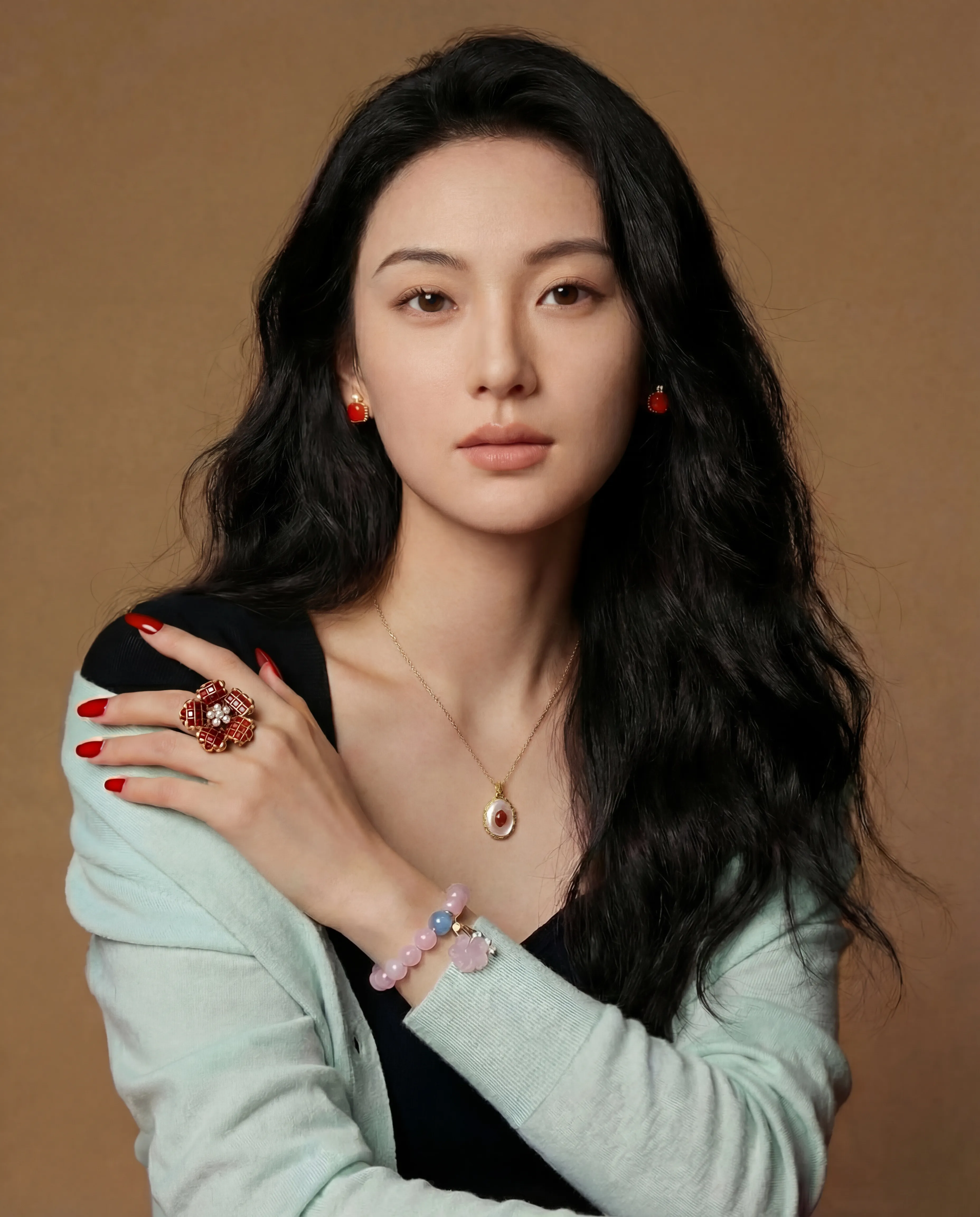 Woman wearing a gold oval pendant necklace, red stone stud earrings, ornate red cocktail ring, and pink beaded bracelet with jade charm.